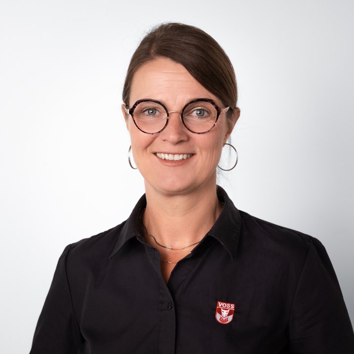 Eine lächelnde Frau mit Brille trägt ein schwarzes Hemd mit einem VOSS-Logo auf der linken Brustseite.