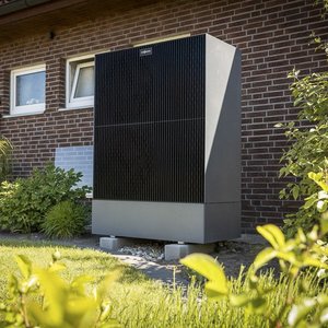 Ein schwarzer, moderner Wärmepumpen-Außenbereich steht neben einem Backsteinhaus, umgeben von grünem Gras und Pflanzen.