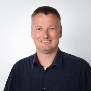 Professionelles Porträt eines lächelnden Mannes mit Hemd Ein lächelnder Mann mit kurzen, grauen Haaren trägt ein dunkelblaues Hemd und steht vor einem hellen Hintergrund.