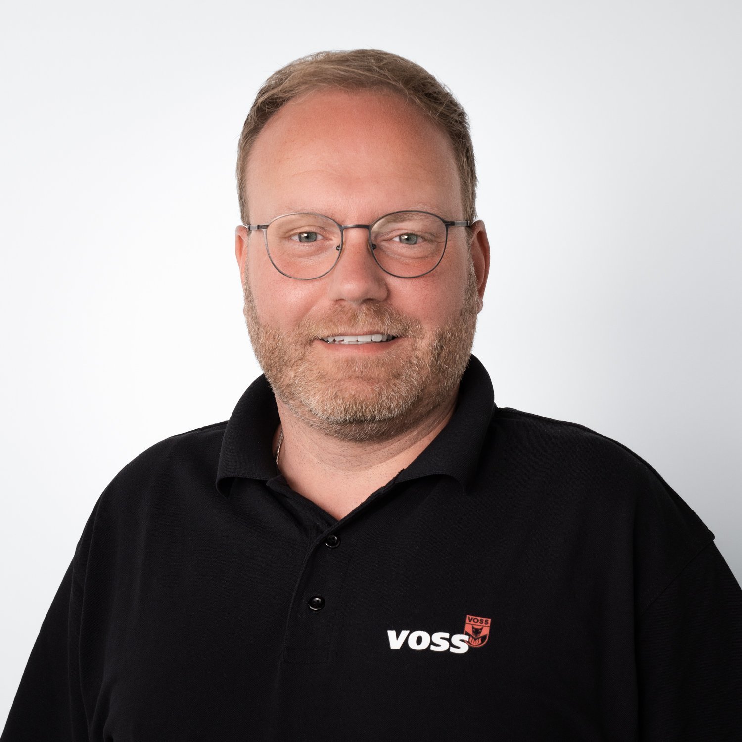 Ein lächelnder Mann mit Brille trägt ein schwarzes Polohemd mit dem Logo von VOSS auf der linken Brustseite.