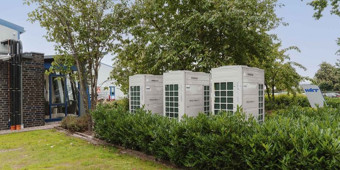 Drei große, weiße Kühlgeräte stehen in einem grünen Gartenbereich, umgeben von Büschen und Bäumen.