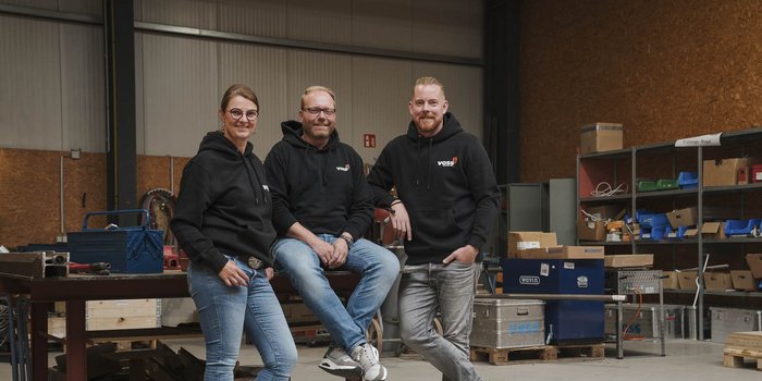 Drei Personen in schwarzen Hoodies stehen lächelnd in einer Werkstatt, umgeben von Werkzeugen und Kisten.