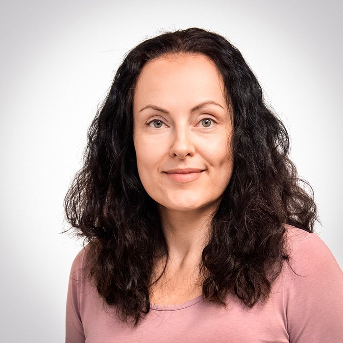 Eine Frau mit lockigem, dunklem Haar lächelt freundlich in die Kamera, während sie ein schlichtes, rosa Oberteil trägt.
