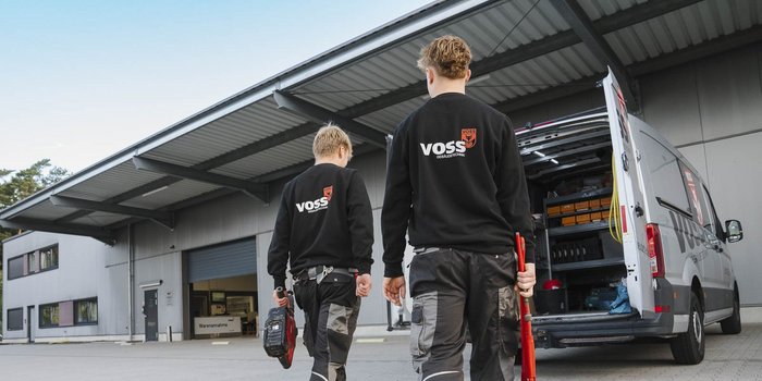 Zwei Techniker in schwarzen Sweatshirts mit dem Logo von VOSS gehen zur Werkstatt, während ein Lieferwagen im Hintergrund steht.