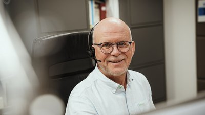 Ein lächelnder Mann mit Brille und Headset sitzt an einem Schreibtisch, umgeben von modernen Büromöbeln.