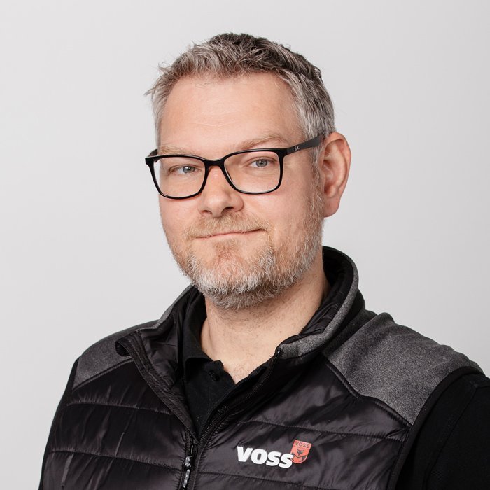 Ein Mann mit Brille und Bart lächelt freundlich in die Kamera, trägt eine schwarze Weste mit dem Logo 'VOSS'.