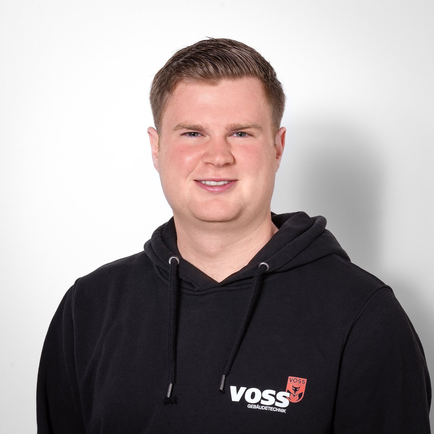 Ein junger Mann mit kurzem, blondem Haar lächelt freundlich in die Kamera, während er einen schwarzen Hoodie trägt.