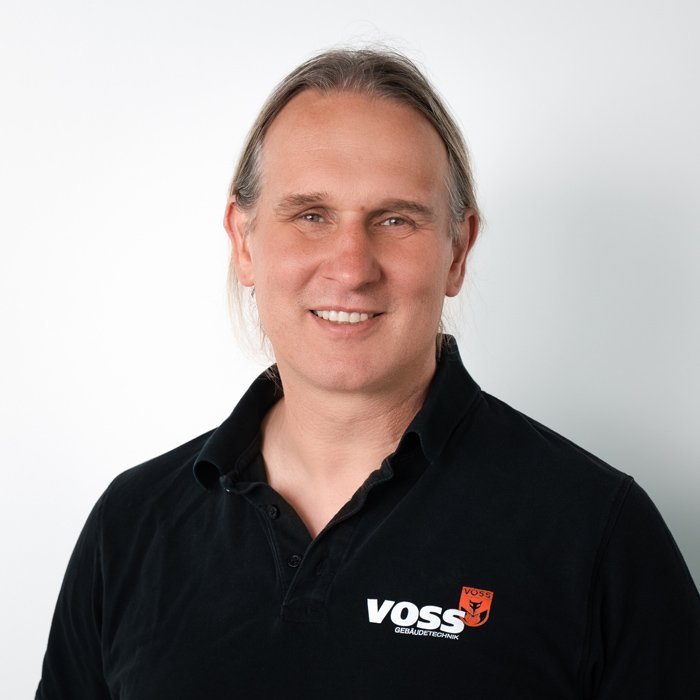 Ein lächelnder Mann mit langen, hellen Haaren trägt ein schwarzes Polohemd mit dem Logo von VOSS auf der linken Brustseite.