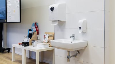 Ein moderner Waschbereich mit einem weißen Waschbecken und einem Handtuchspender an der Wand, umgeben von hellen Fliesen.