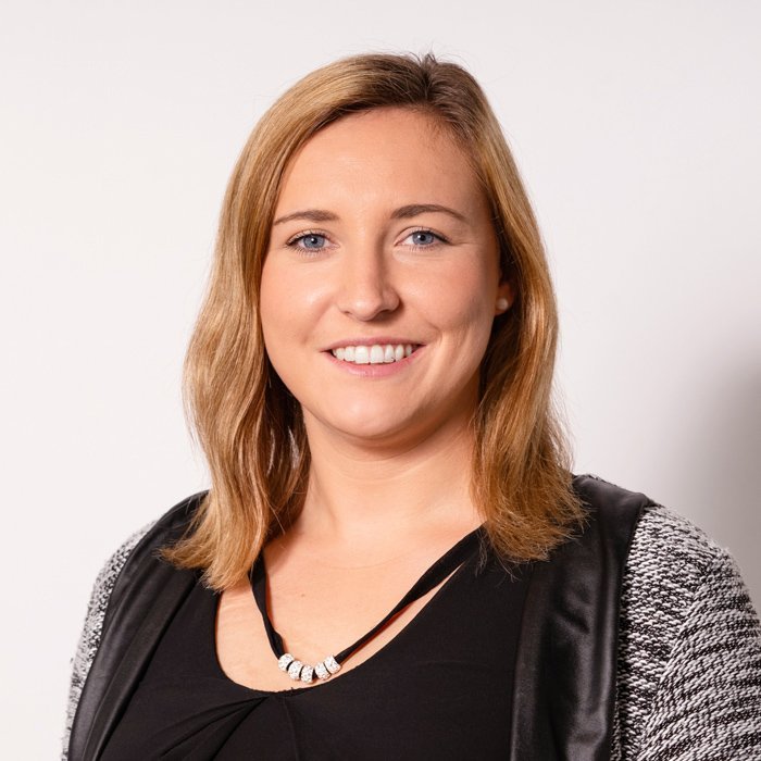 Eine lächelnde Frau mit blonden Haaren trägt eine schwarze Bluse und eine auffällige Halskette vor einem neutralen Hintergrund.