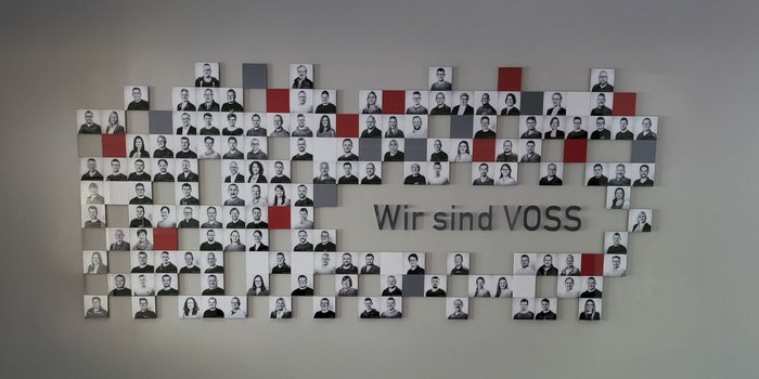Eine Wand mit vielen schwarz-weißen Porträts von Mitarbeitern, die das Motto 'Wir sind VOSS' zeigt.