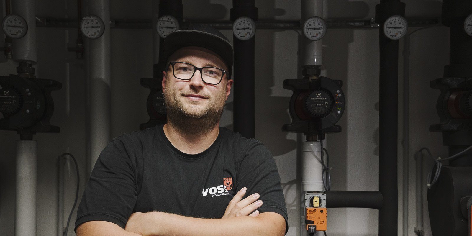 Ein junger Mann mit Brille und schwarzem T-Shirt steht lächelnd vor einer Reihe von Rohren und Druckmessgeräten.