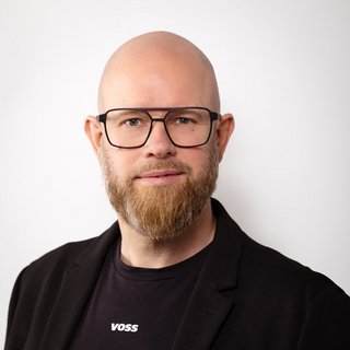Professionelles Porträt eines Mannes mit Bart und Brille Ein Mann mit Glatze und Bart trägt eine schwarze Jacke und ein T-Shirt mit dem Schriftzug 'VOSS', lächelt freundlich.