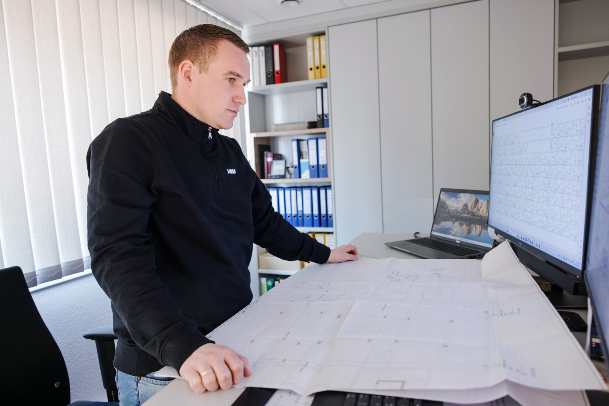 Ein Mann steht an einem Schreibtisch und betrachtet sorgfältig Baupläne, während er an einem Computer arbeitet.