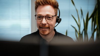 Ein lächelnder Mann mit Brille und Headset sitzt vor einem Computerbildschirm in einem hellen Büro.