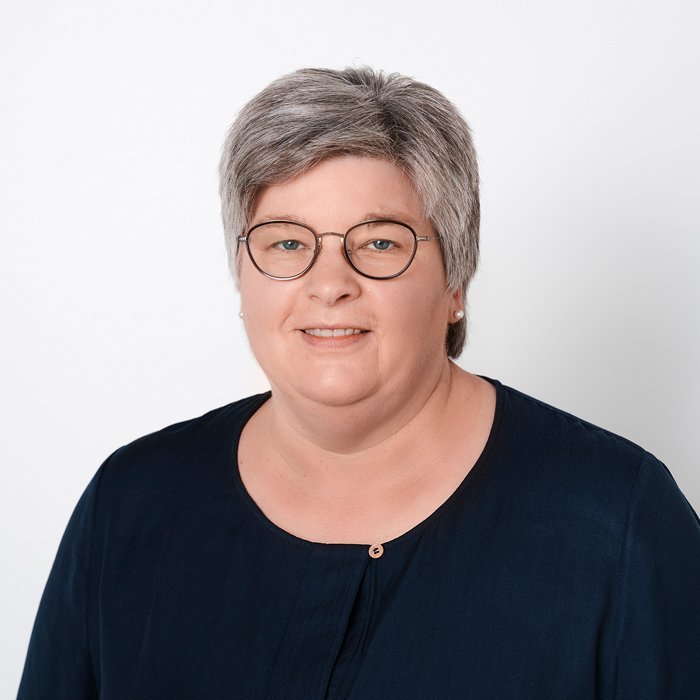 Eine Frau mit grauen Haaren und Brille lächelt freundlich vor einem weißen Hintergrund, trägt ein dunkelblaues Oberteil.