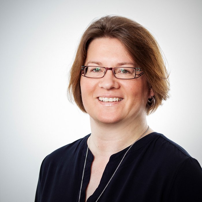 Eine lächelnde Frau mit Brille und kurzen, braunen Haaren trägt ein dunkles Oberteil und steht vor einem hellen Hintergrund.