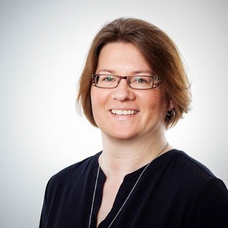 Helle Studioaufnahme einer Frau mit freundlichem Ausdruck Eine lächelnde Frau mit Brille und kurzen, braunen Haaren trägt ein dunkles Oberteil und steht vor einem hellen Hintergrund.