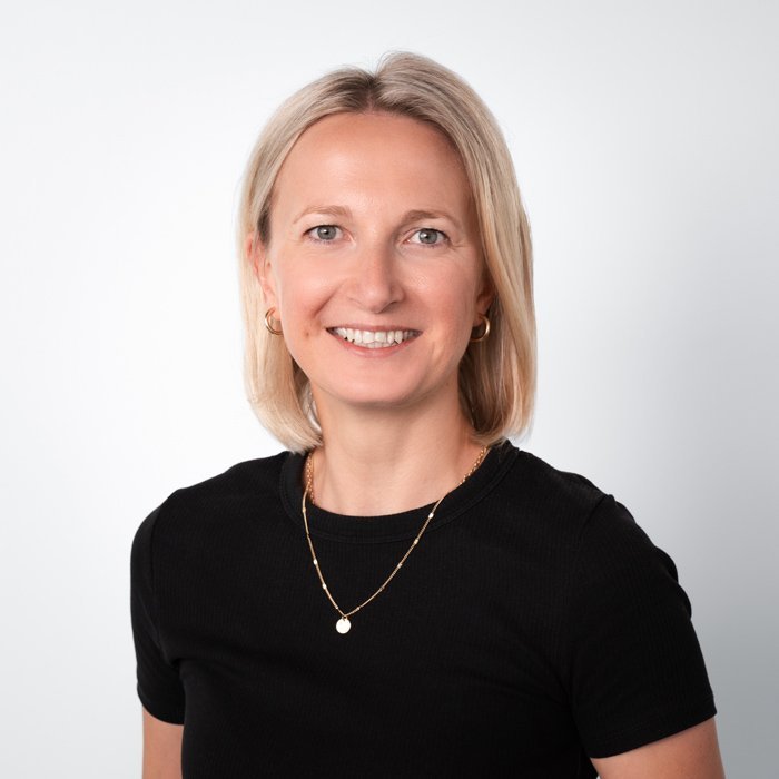 Eine lächelnde Frau mit blonden Haaren trägt ein schwarzes T-Shirt und eine goldene Halskette vor einem hellen Hintergrund.