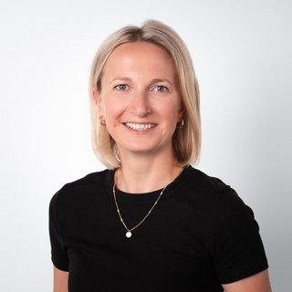 Professionelles Porträt einer lächelnden Frau mit blonden Haaren Eine lächelnde Frau mit blonden Haaren trägt ein schwarzes T-Shirt und eine goldene Halskette vor einem hellen Hintergrund.