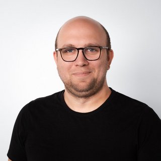 Porträt eines lächelnden Mannes mit Brille Ein Mann mit Brille und Glatze lächelt freundlich in die Kamera, trägt ein schwarzes T-Shirt vor einem hellen Hintergrund.