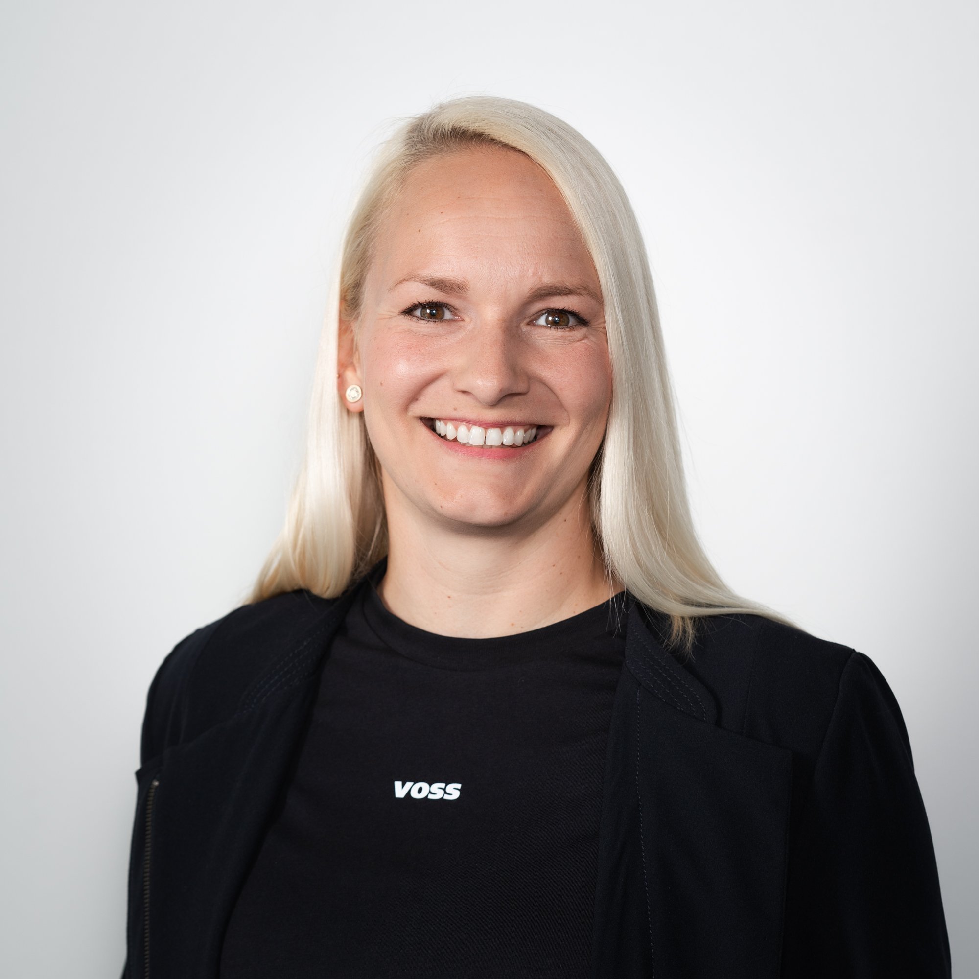 Eine lächelnde Frau mit langen, blonden Haaren trägt ein schwarzes Oberteil mit dem Schriftzug 'VOSS' und eine Jacke.