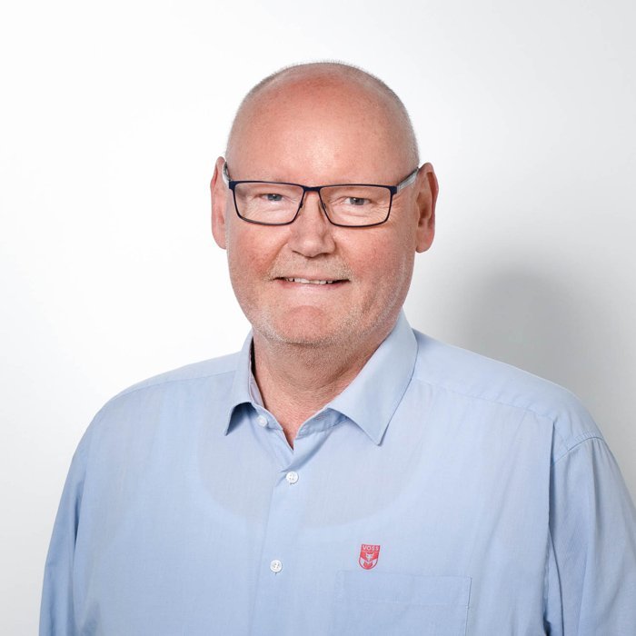 Ein lächelnder Mann mit Glatze trägt eine hellblaue Hemd und eine Brille, vor einem weißen Hintergrund.