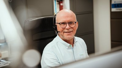 Ein lächelnder Mann mit Brille und Headset sitzt an einem Schreibtisch in einem modernen Büro.