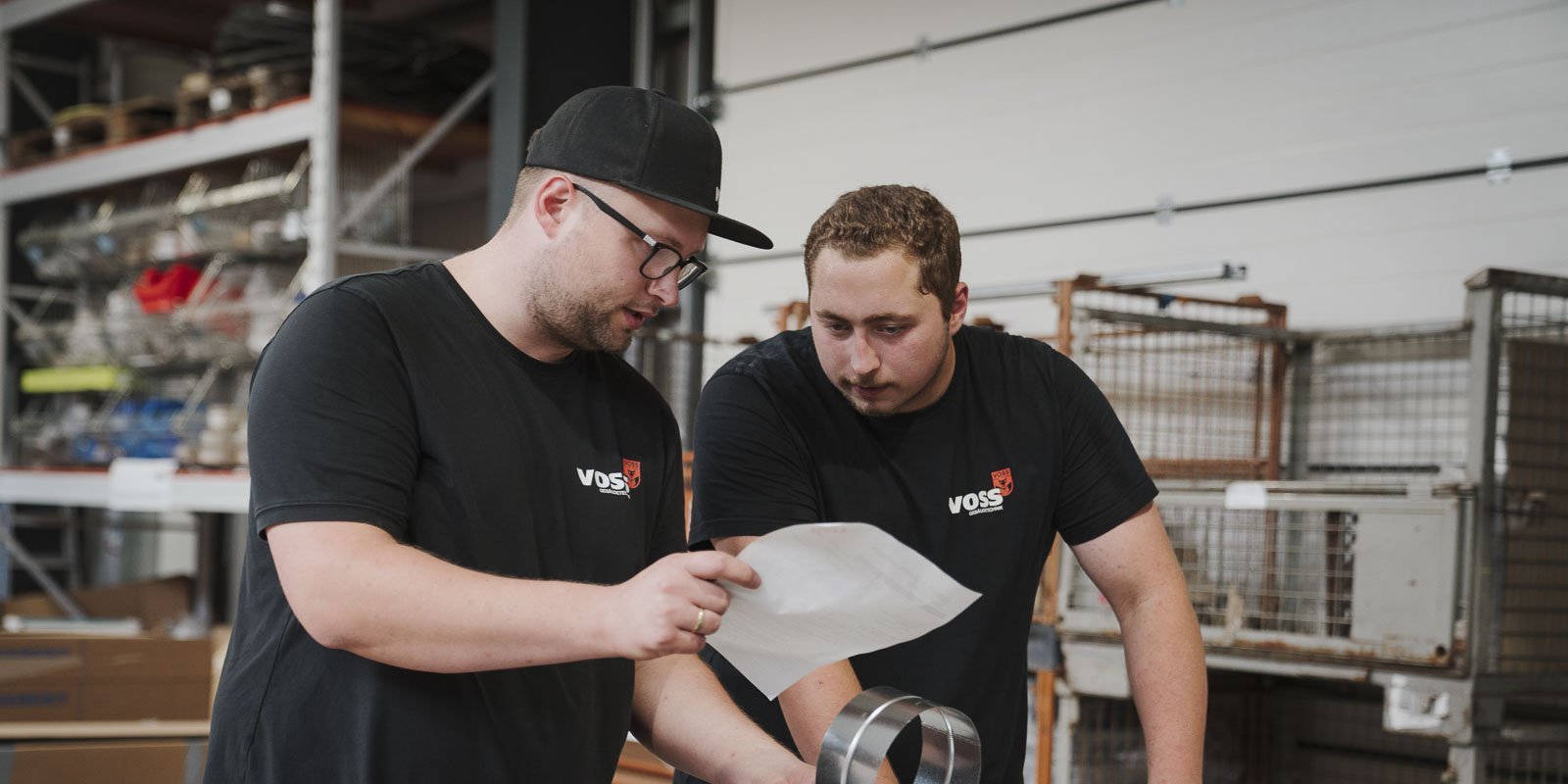 Zwei Männer in schwarzen T-Shirts stehen in einer Werkstatt und studieren ein Dokument, während sie eine Metallring halten.