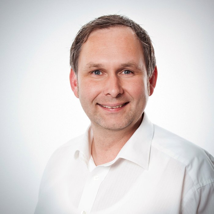 Ein lächelnder Mann mit kurzen, braunen Haaren trägt ein weißes Hemd und steht vor einem hellen Hintergrund.