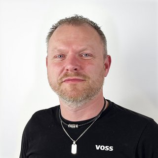Porträt eines Mannes mit Bart und Halskette Ein Mann mit kurzem, grauem Haar und Bart trägt ein schwarzes T-Shirt mit dem Aufdruck 'VOSS' und mehrere Halsketten.