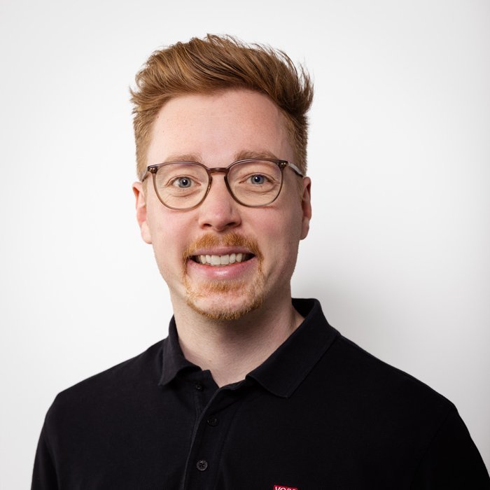 Ein junger Mann mit Brille und einem freundlichen Lächeln trägt ein schwarzes Polohemd vor einem weißen Hintergrund.