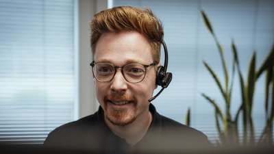 Ein lächelnder Mann mit Brille und Headset sitzt vor einem Computer, umgeben von Pflanzen und Fensterjalousien.