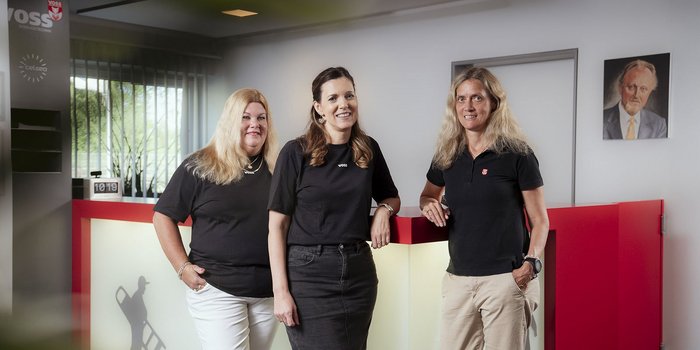 Drei Frauen in schwarzen T-Shirts stehen lächelnd an einem Empfangstresen, umgeben von modernen Büromöbeln und Pflanzen.