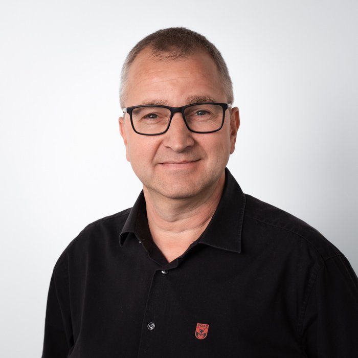Ein Mann mit Brille lächelt freundlich in die Kamera, trägt ein schwarzes Hemd mit einem kleinen Logo auf der Brust.