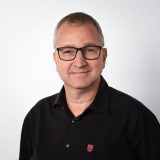 Professionelles Porträt eines lächelnden Mannes in Brille Ein Mann mit Brille lächelt freundlich in die Kamera, trägt ein schwarzes Hemd mit einem kleinen Logo auf der Brust.