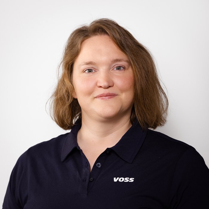 Eine Frau mit schulterlangen, braunen Haaren lächelt freundlich in die Kamera, trägt ein dunkelblaues Poloshirt mit dem Logo VOSS.