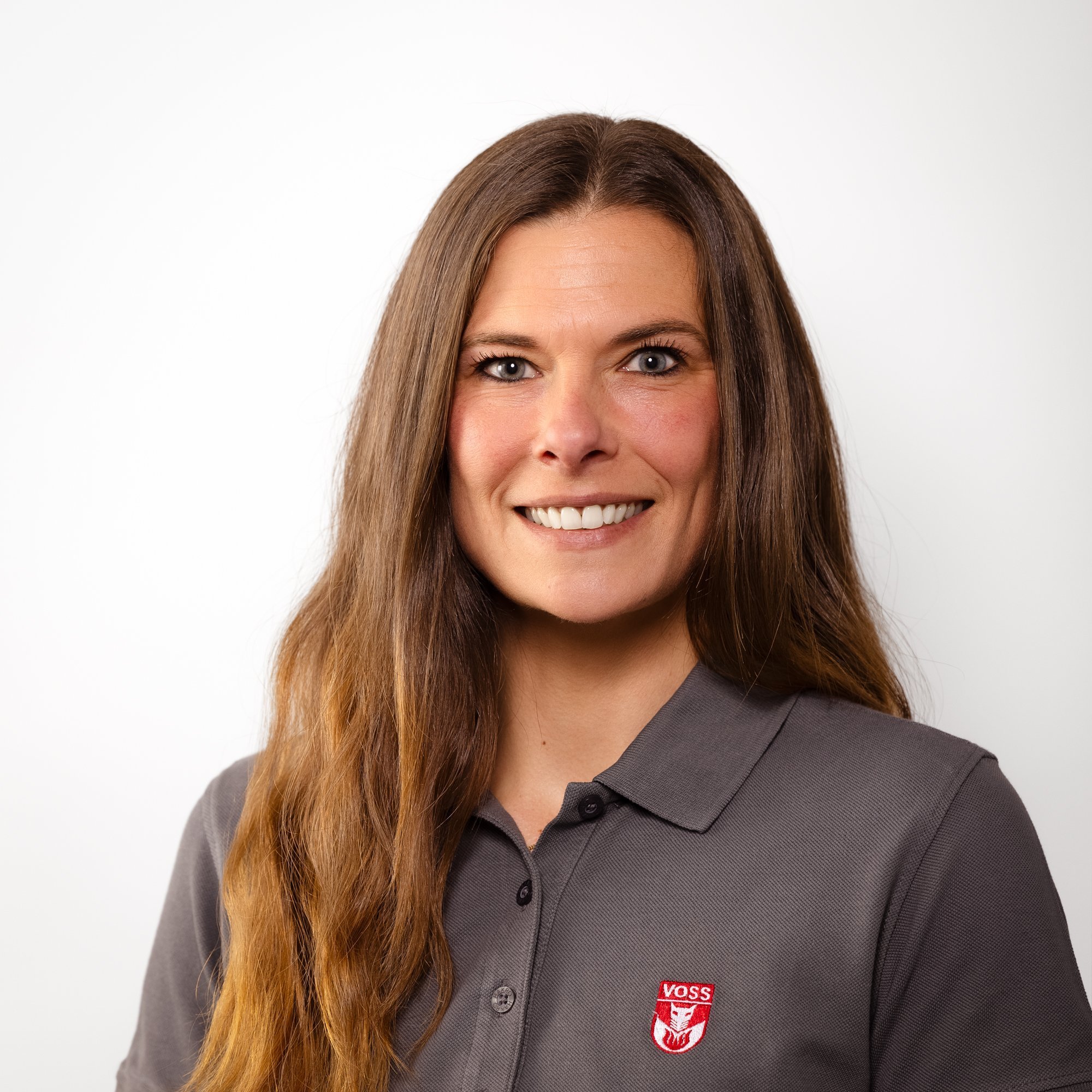 Eine Frau mit langen, braunen Haaren lächelt freundlich in einem grauen Poloshirt mit dem Logo von VOSS.