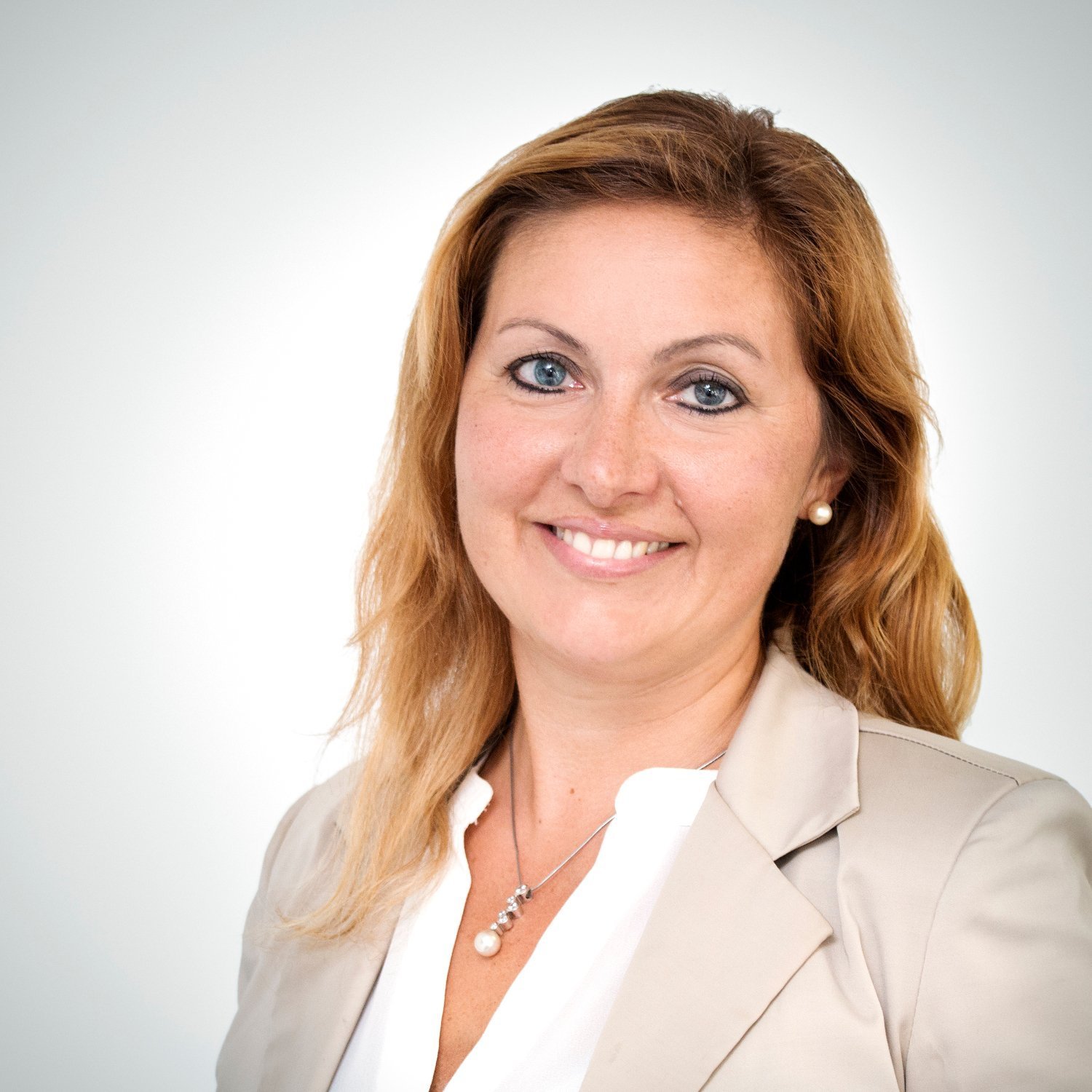 Eine lächelnde Frau mit blonden Haaren trägt einen hellen Blazer und eine Perlenkette, vor einem hellen Hintergrund.