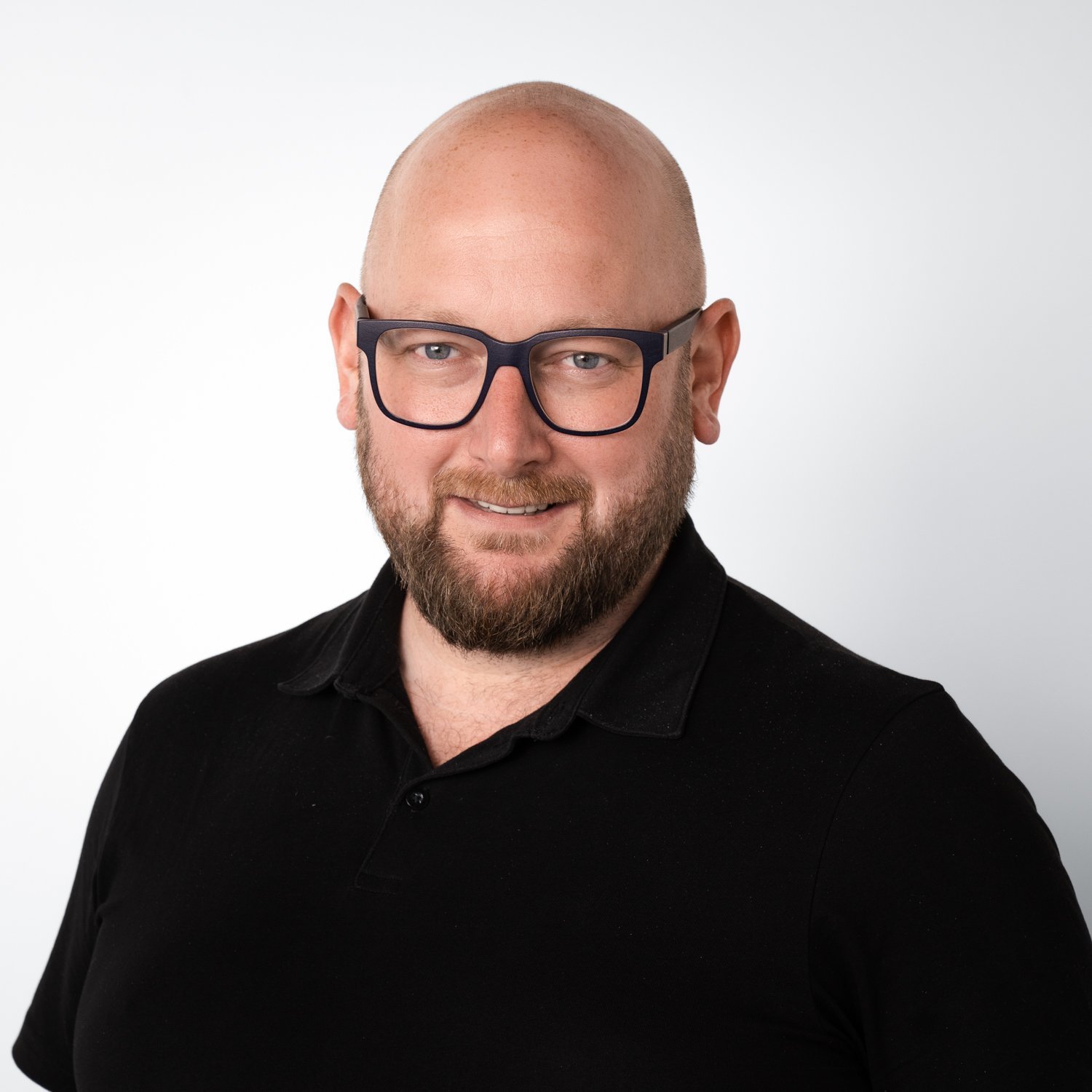 Ein lächelnder Mann mit Glatze und Bart trägt eine schwarze Polohemd und eine modische Brille vor einem hellen Hintergrund.
