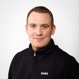 Professionelles Foto eines Mannes mit neutralem Hintergrund Ein junger Mann mit kurzem, braunem Haar lächelt freundlich in die Kamera, während er einen schwarzen Reißverschlusspullover trägt.