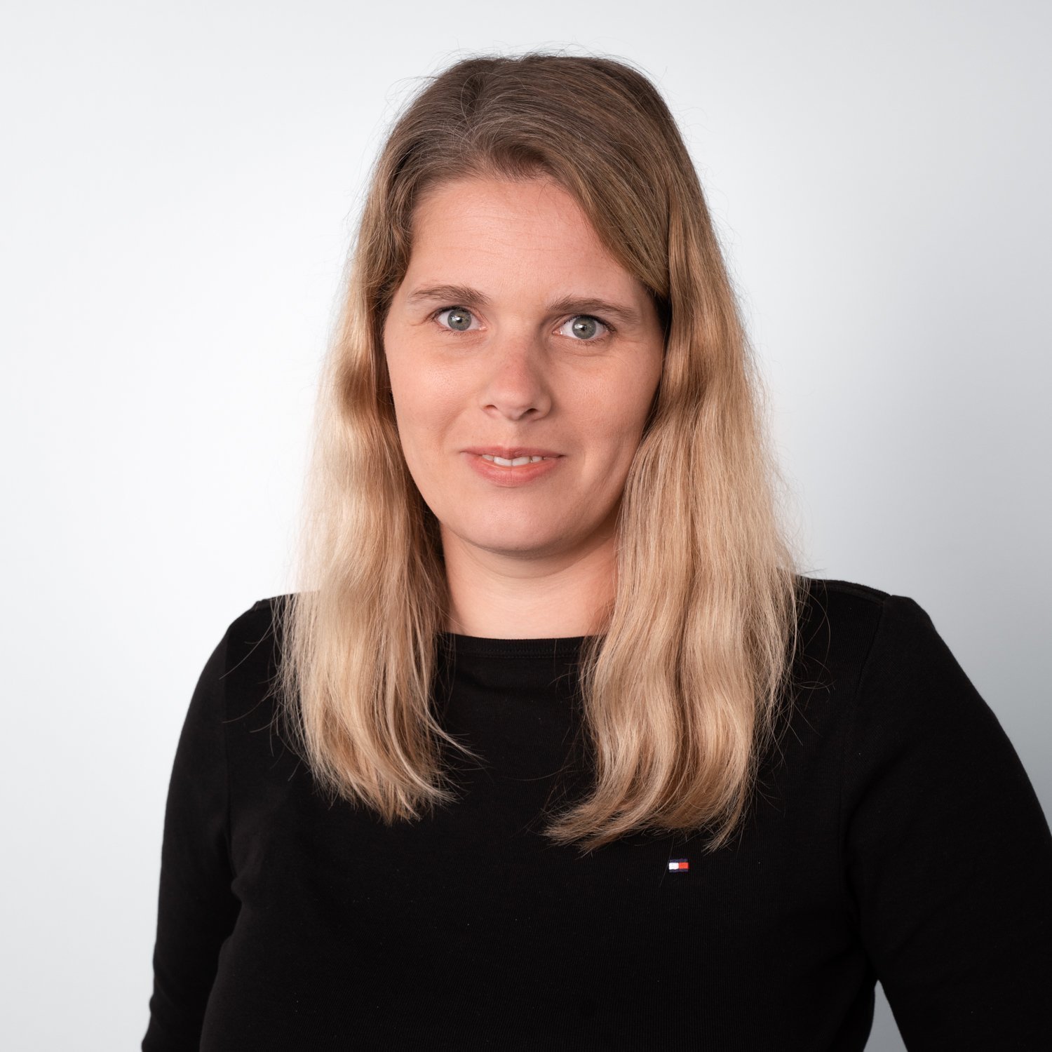 Eine Frau mit langen, blonden Haaren lächelt freundlich in die Kamera, während sie ein schwarzes Oberteil trägt.
