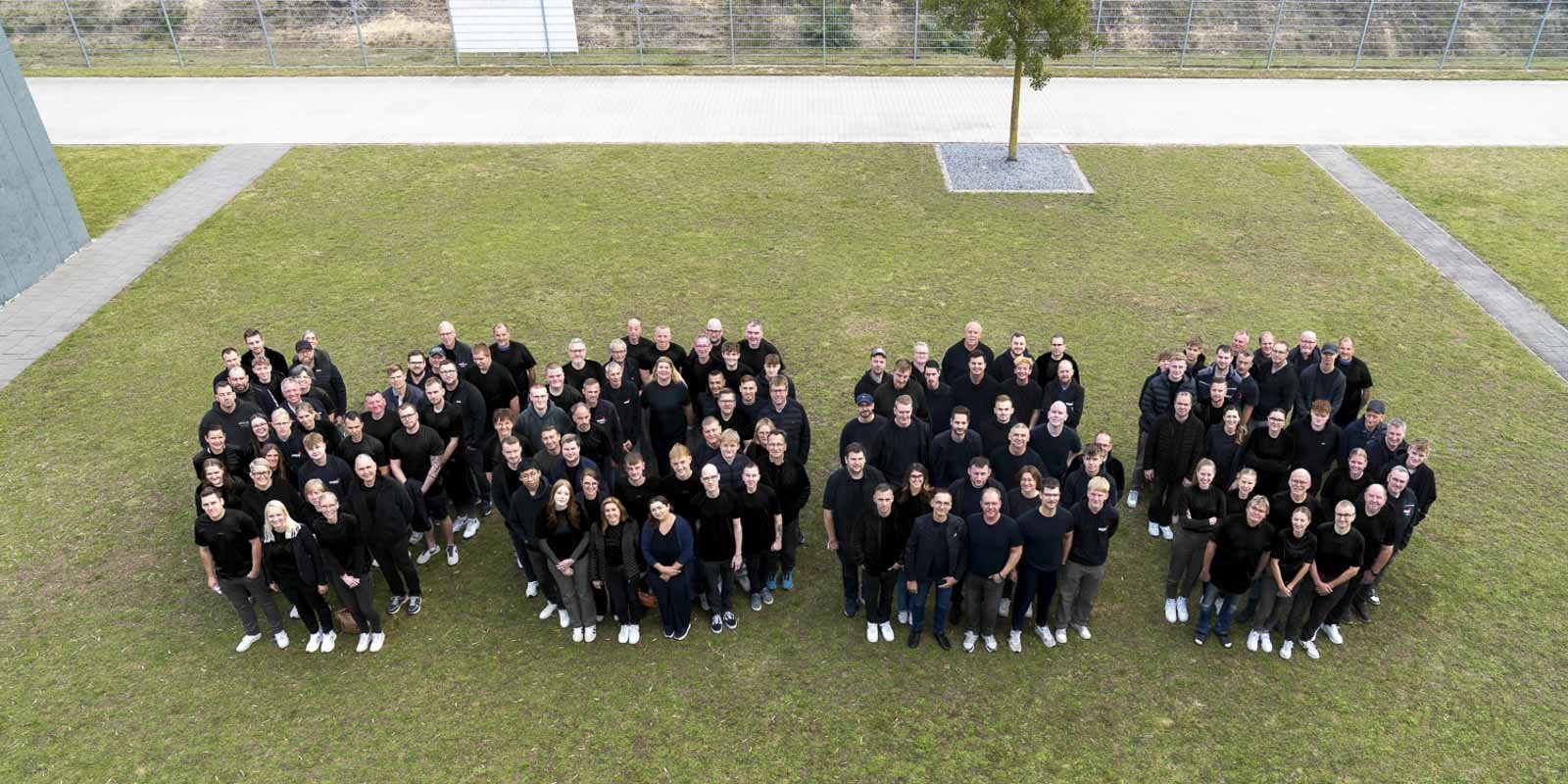 Eine große Gruppe von Menschen steht auf einer Wiese und trägt schwarze Kleidung, um ein gemeinsames Bild zu schaffen.