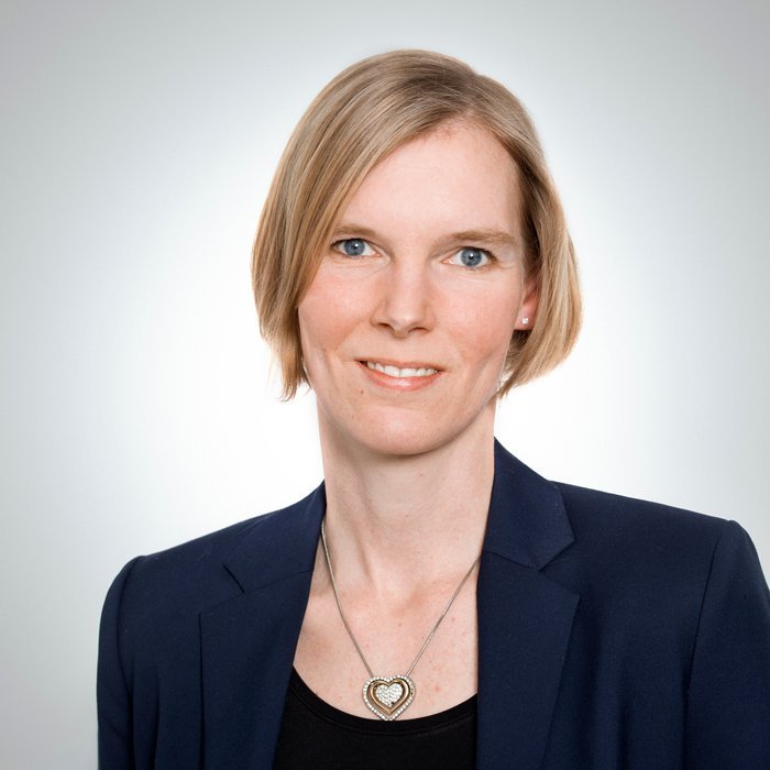 Eine Frau mit blonden, schulterlangen Haaren lächelt freundlich in die Kamera, trägt einen blauen Blazer und eine Herz-Halskette.