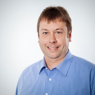 Professionelles Porträt eines lächelnden Mannes in Hemd Ein Mann mit kurzen, braunen Haaren lächelt freundlich in die Kamera, gekleidet in ein hellblaues Hemd.