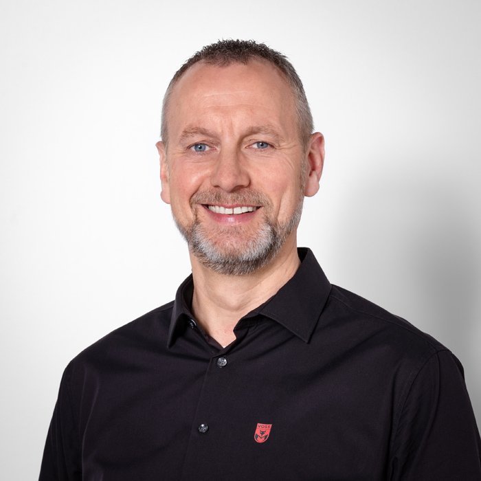 Ein lächelnder Mann mit grauem Bart trägt ein schwarzes Hemd und steht vor einem hellen Hintergrund.