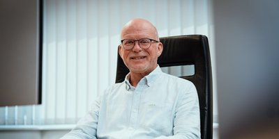 Ein älterer Mann mit Brille lächelt freundlich in einem modernen Büro, umgeben von hellen Vorhängen und einem eleganten Stuhl.