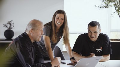 Drei Personen in einem modernen Büro diskutieren lebhaft über einen Laptop, während eine Pflanze im Vordergrund sichtbar ist.
