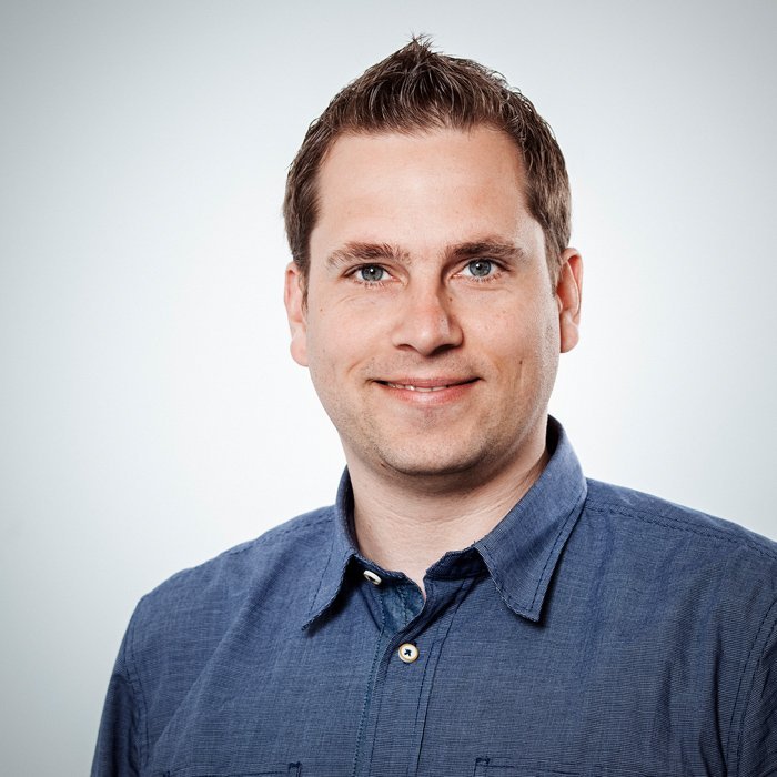 Ein junger Mann mit kurzen, gestylten Haaren lächelt freundlich in die Kamera, während er ein blaues Hemd trägt.
