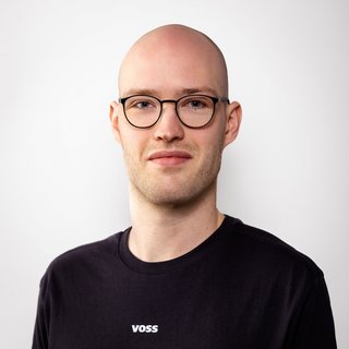 Porträt eines jungen Mannes mit Brille und Glatze Ein junger Mann mit Glatze und Brille lächelt leicht vor einem hellen Hintergrund, trägt ein schwarzes T-Shirt mit dem Schriftzug 'VOSS'.
